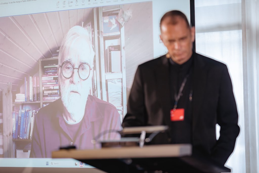 A man in glasses with white hair and a beard appears on a large video call screen, sitting in front of bookshelves. In the foreground, another man stands at a lectern, slightly blurred, facing the audience with a red badge visible on his chest.