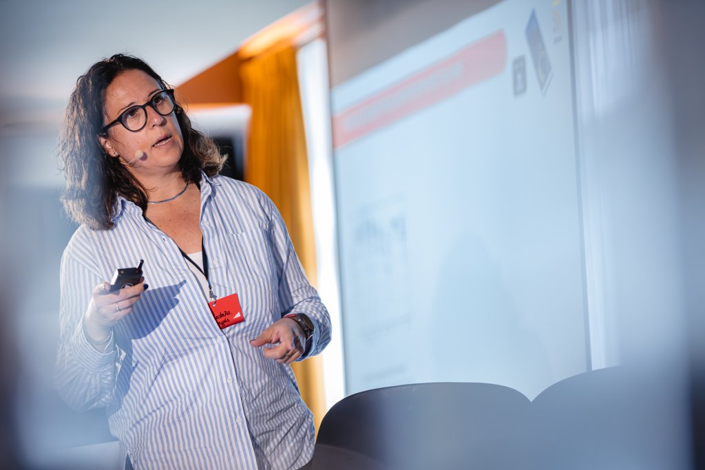 A woman wearing glasses, a striped shirt, and a headset holds a remote whilst presenting in front of a projected screen. She has dark hair and a red badge around her neck. The background is softly lit with a yellow curtain visible.