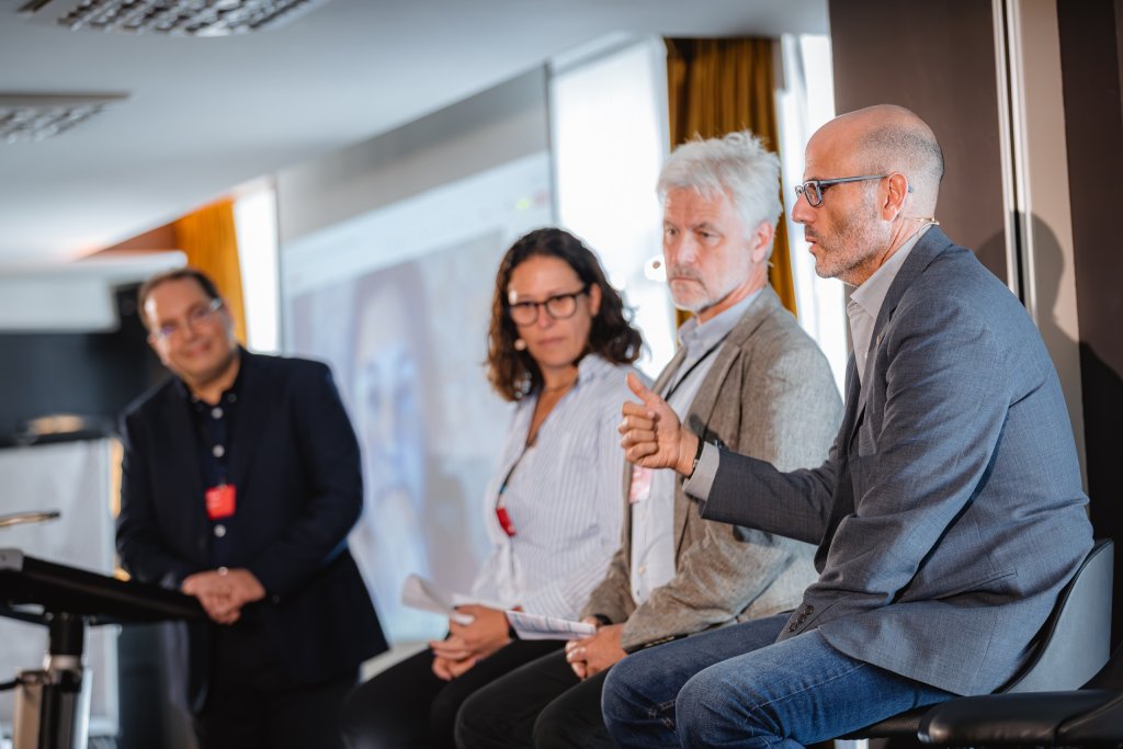 Four people are engaged in a panel discussion. Three sit on high stools, listening as a bald man in glasses and a blazer speaks, gesturing with his hand. A fourth person stands to the side, smiling. A screen with a video call is visible in the background.