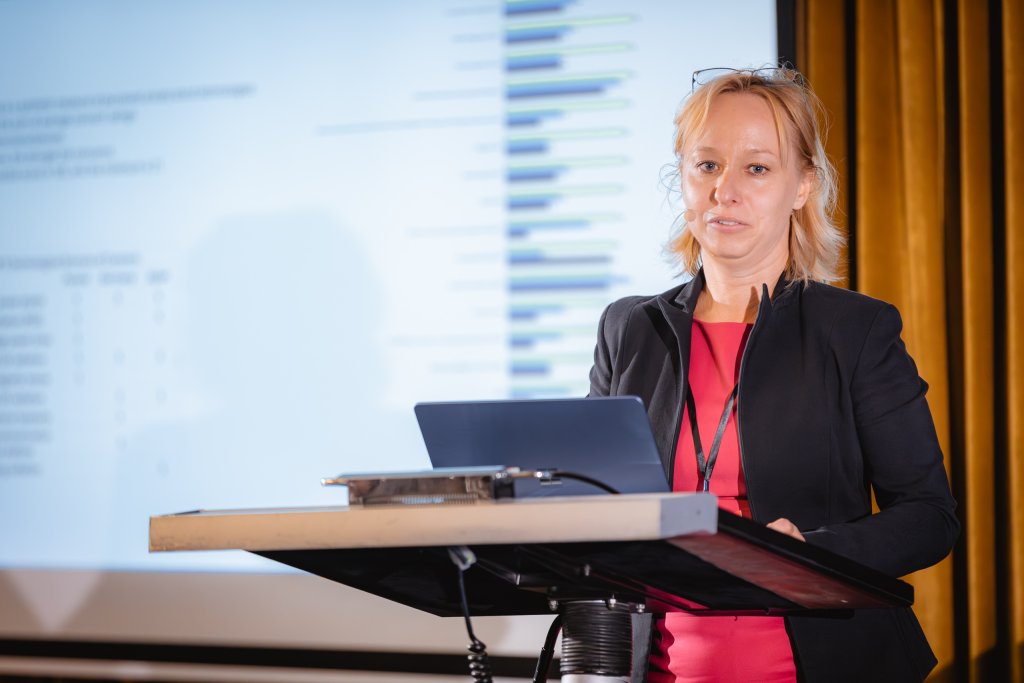 A woman with blonde hair, wearing a black blazer over a red top, stands at a lectern with a laptop in front of her. She is speaking, with a large projected graph and blurred text visible on a screen behind her, and yellow curtains to the side.