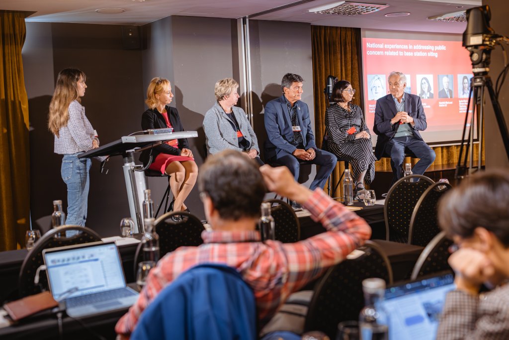 A panel of five professionals—three men and two women—sits on stage discussing a topic, while a woman moderator stands with a microphone. Audience members sit in front, some using laptops. A presentation slide is projected behind the panellists.
