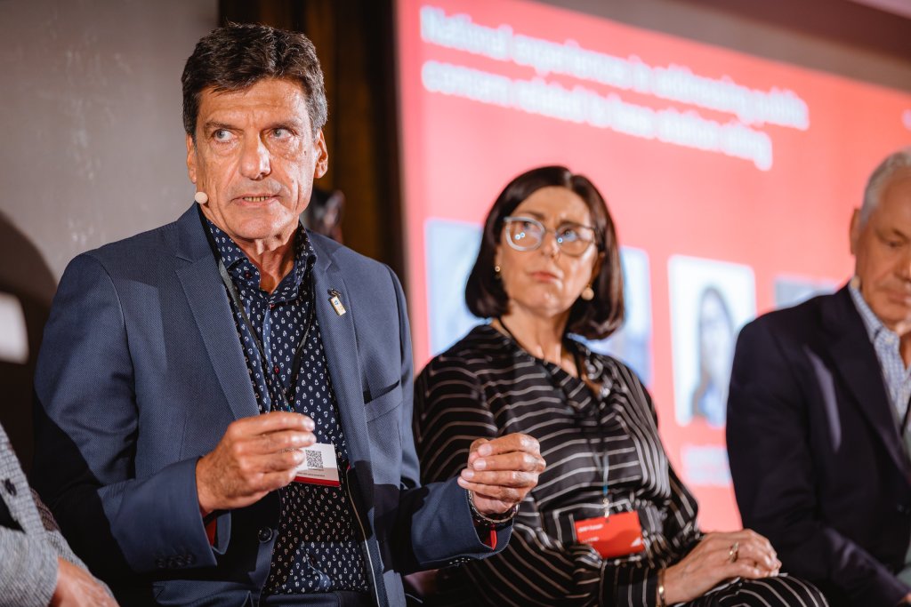 A man in a blue suit speaks while holding an object, seated next to a woman in glasses and a striped blouse, and another man. They are on a stage with red name badges and a blurred red presentation screen in the background.