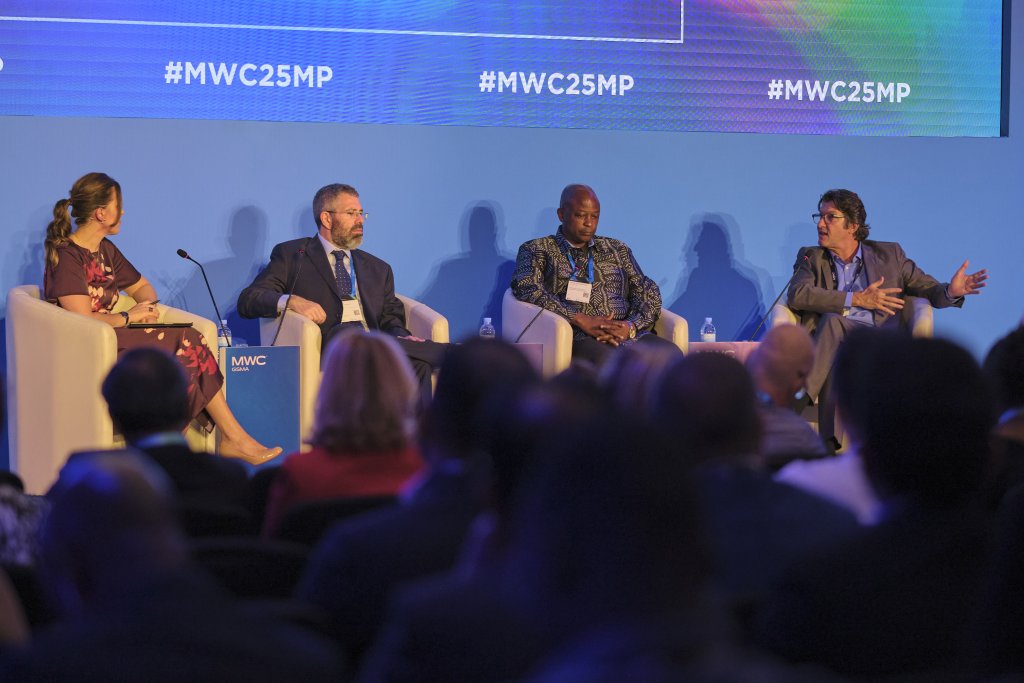 Four panellists, three men and one woman, sit on stage in white chairs, engaged in discussion at a conference. The background is blue with "#MWC25MP" displayed multiple times. The audience is visible in the foreground, listening attentively.