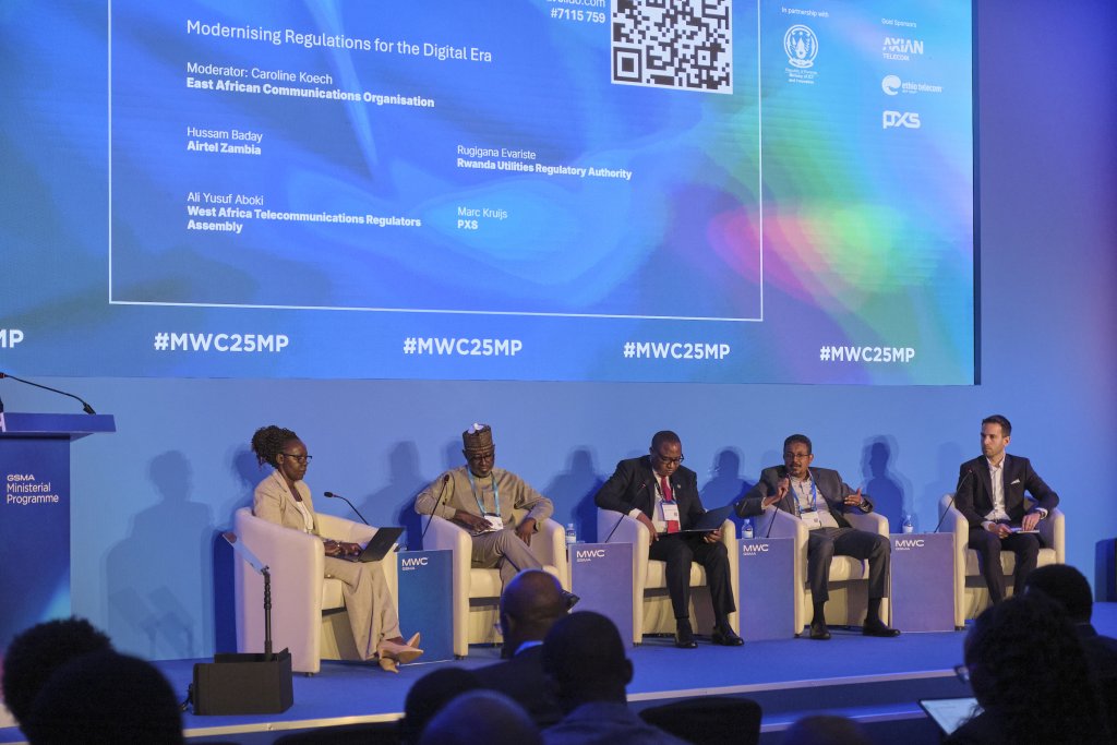 Five panellists sit on a stage with blue lighting and a digital screen behind them displaying the session title, speakers' names, and hashtags. They are engaged in a discussion on modernising digital regulations. The audience is visible in the foreground.