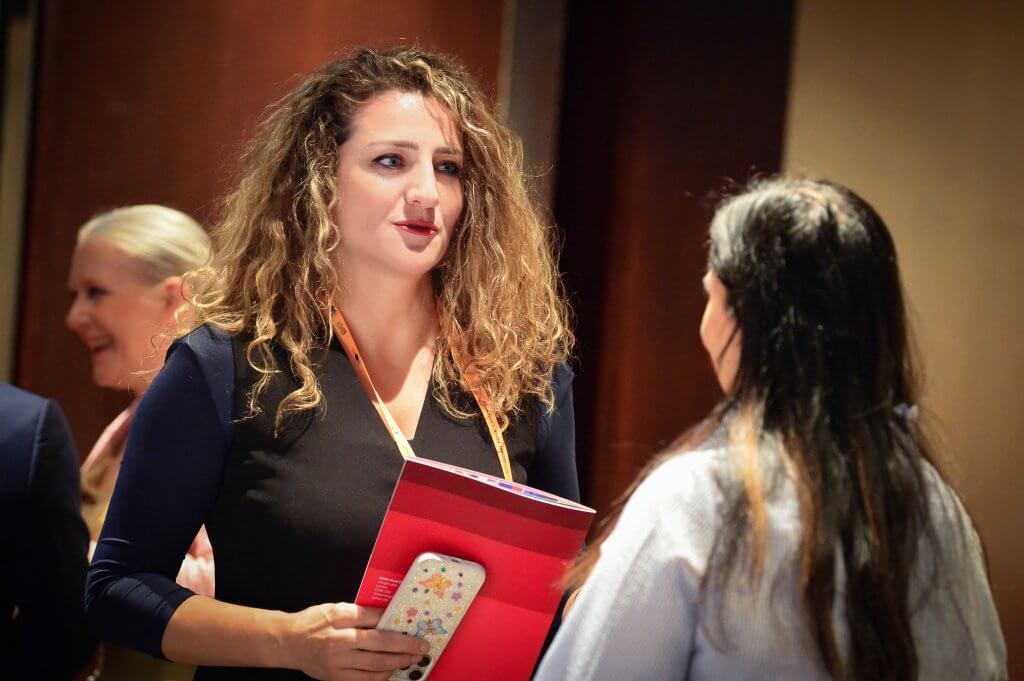 A woman with curly hair, wearing a dark dress, holds a colourful phone and a red folder whilst talking to another woman with long dark hair. They are indoors at an event, with another person blurred in the background.