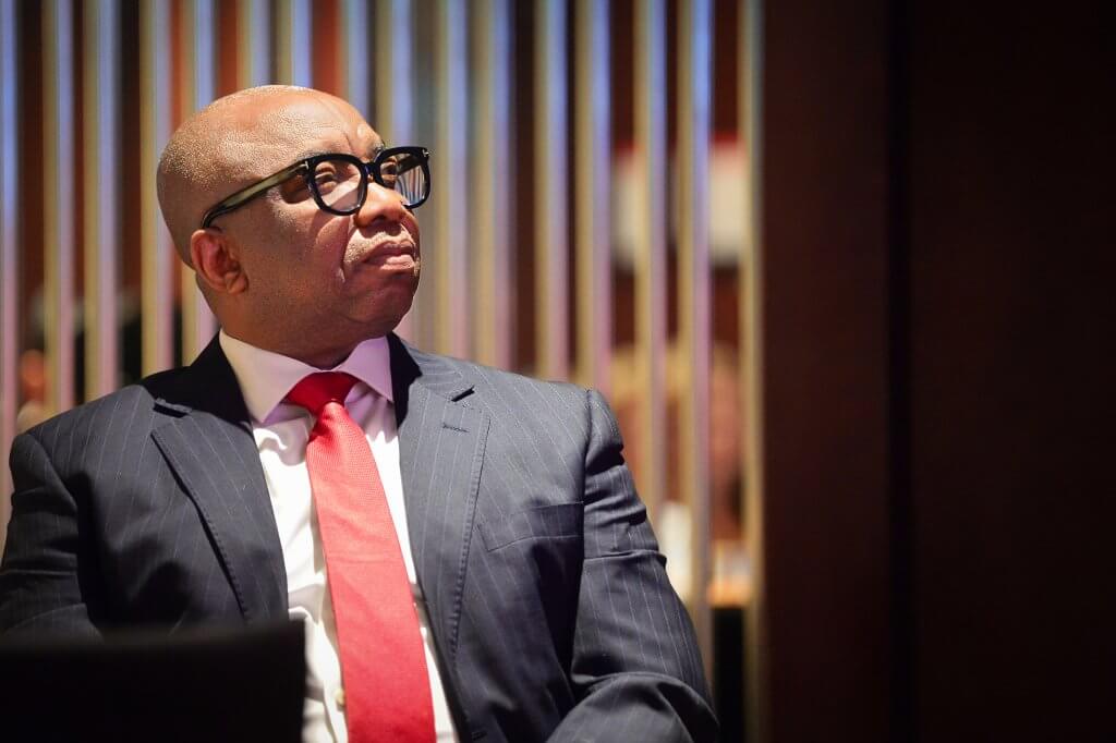 A man in a blue pinstripe suit, white shirt, and red tie sits looking upward thoughtfully. He wears black-rimmed glasses and is bald. The background is softly blurred with vertical metallic bars and warm lighting.