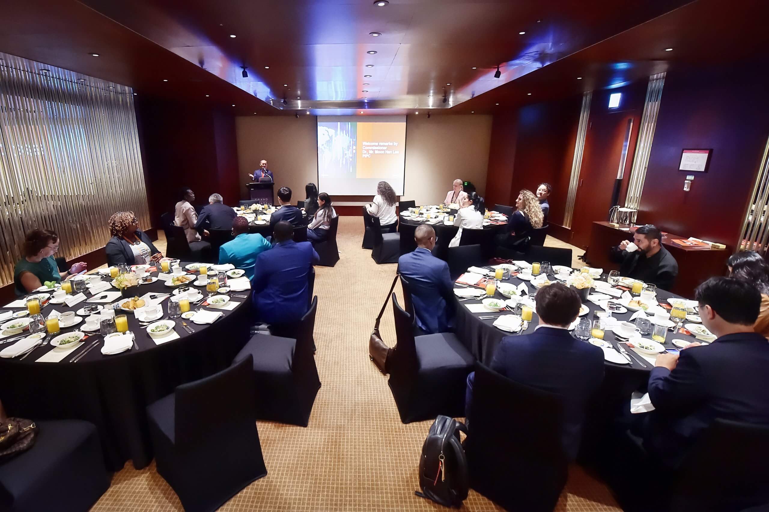 A conference room with round tables, each seating several people in business attire. A speaker stands at a lectern by a screen presenting slides. Plates of food and drinks are on tables. Attendees listen attentively in a warmly lit, modern setting.