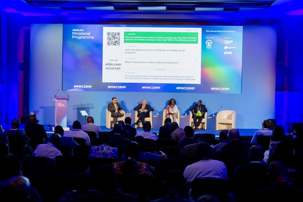 A panel of four people sits on a stage in front of a large screen displaying a question at a conference. The audience faces the stage. The event backdrop includes logos and blue lights, with a lectern on the left side.