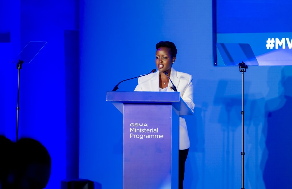A woman in a white blazer stands at a podium labelled "GSMA Ministerial Programme," speaking into two microphones. Two transparent autocues are positioned on either side. The background is blue, with a screen partially showing a hashtag.