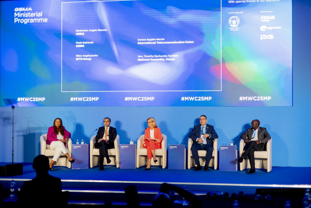 Five panellists, three men and two women, sit on a brightly lit stage with a blue backdrop displaying names and organisations. They are each seated in white chairs with microphones and water bottles in front. The event branding reads "#MWC25MP" and "GSMA Ministerial Programme.