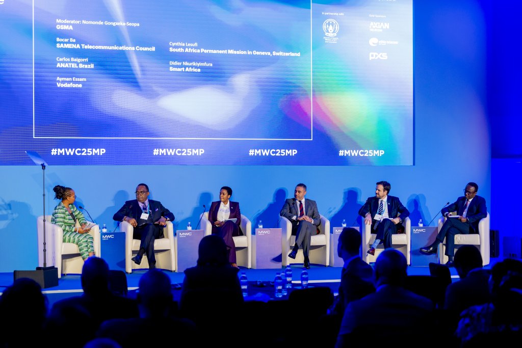 Five panellists—three men and two women—sit on a stage discussing at MWC25. Each has a microphone and is seated in white armchairs. Behind them, a large blue screen displays speaker names and hashtags. Audience silhouettes are visible in the foreground.