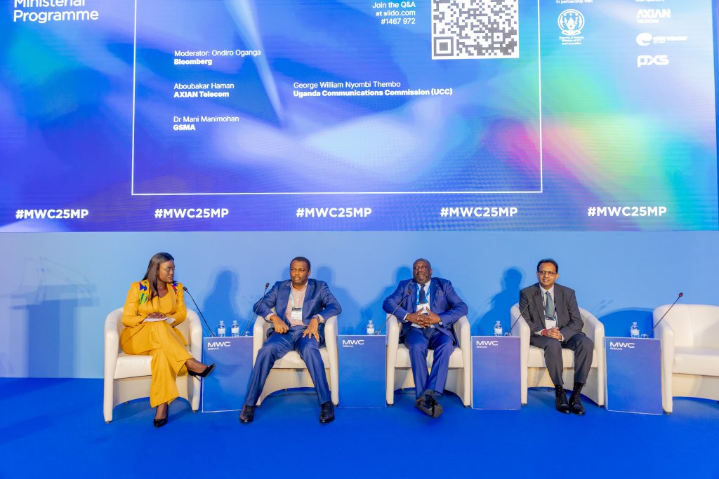 Four panellists sit on stage in armchairs against a blue backdrop with event logos and a QR code. The woman on the left wears a yellow trouser suit, the three men wear suits. White tables and water bottles are in front of them. #MWC25MP is visible.