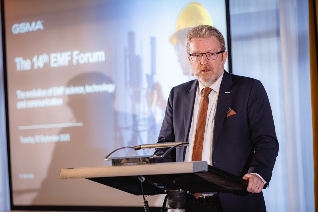 A bearded man in a suit and glasses speaks at a podium. Behind him, a large screen displays "GSMA The 14th EMF Forum" with EMF Forum 2025 takeaways, alongside industrial imagery and a person in a yellow hard hat.