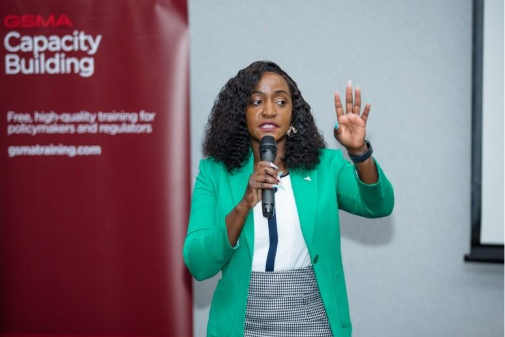 A woman with curly hair in a green blazer speaks into a microphone and raises her hand, gesturing during a presentation. Behind her is a red banner with “GSMA Capacity Building” and text about training for policymakers and regulators.