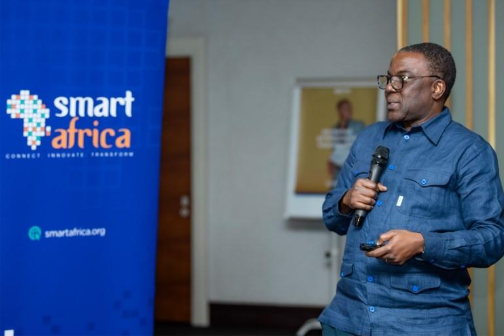 A man in glasses and a blue shirt holds a microphone, speaking at an event. To his left, a blue banner displays the Smart Africa logo and the words "Connect Innovate Transform." A blurred poster and door are visible in the background.