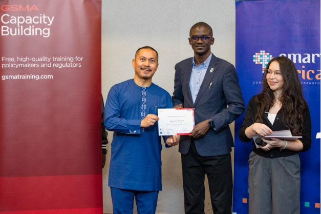 Three people stand indoors; one man in blue traditional attire holds a certificate and smiles at another man in a suit who is presenting it. A woman with papers stands beside them. Banners for GSMA and Smart Africa highlight mobile privacy and security in the background.