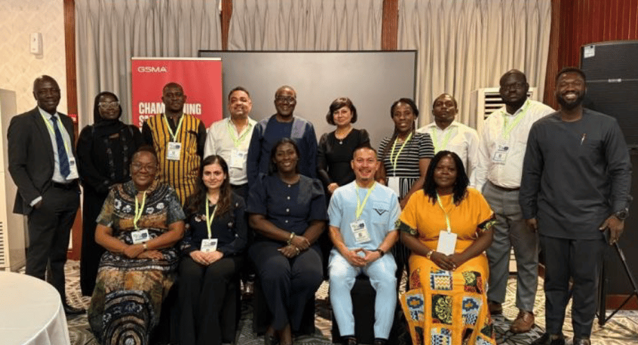 A group of fifteen people, both men and women, pose and smile in two rows indoors. Most wear business or business-casual attire with conference lanyards. Behind them are curtains, a red “GSMA” banner, and a projection screen highlighting smart data privacy.