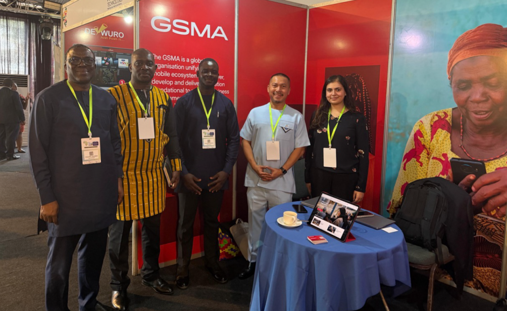 Five people stand and smile for a photo at a GSMA exhibition stand, highlighting smart data privacy solutions. Three men wear traditional West African attire, one man wears a light blue outfit, and one woman wears black. A round table displays a cup, saucer, and tablet.