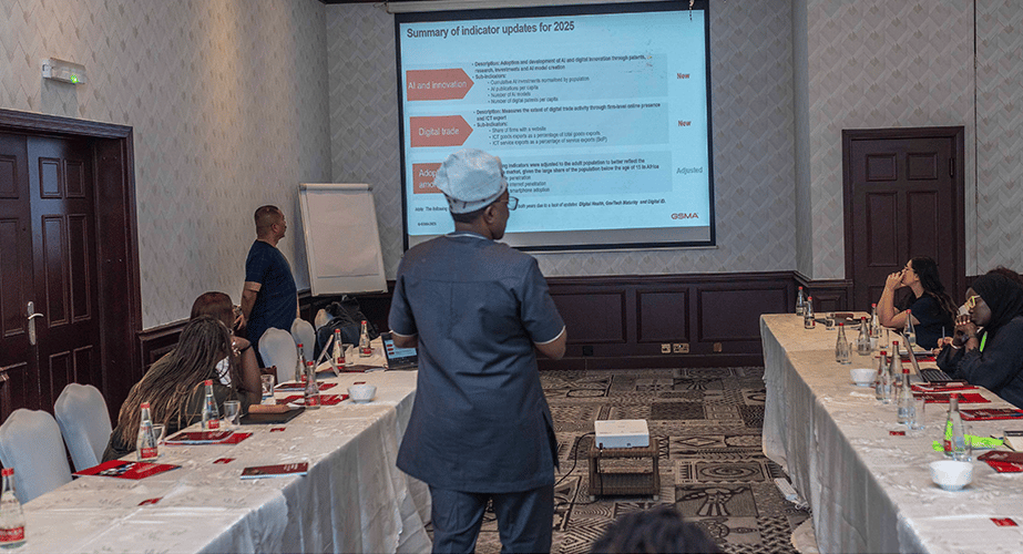 A group of people sit around a U-shaped table in a conference room, listening to a presenter in grey discuss smart data privacy. A projector screen displays 2025 indicator updates, whilst another stands near a whiteboard with bottles and papers on the tables.