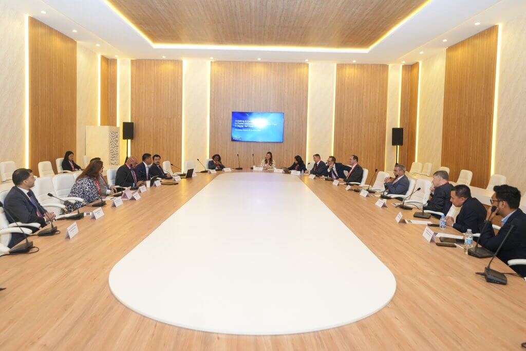 A group of people in business attire sit around a large white oval conference table in a spacious, modern room with wood-panelled walls. A presentation screen is on the far wall, and nameplates are on the table in front of each participant.