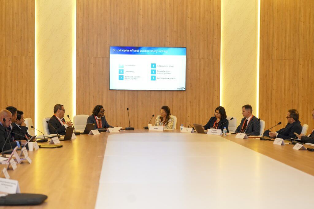 A group of professionals in business attire sit around a large, oval conference table in a modern boardroom with wood-panelled walls. A screen displays “Six principles of best critical policy making.” Nameplates and microphones are visible on the table.