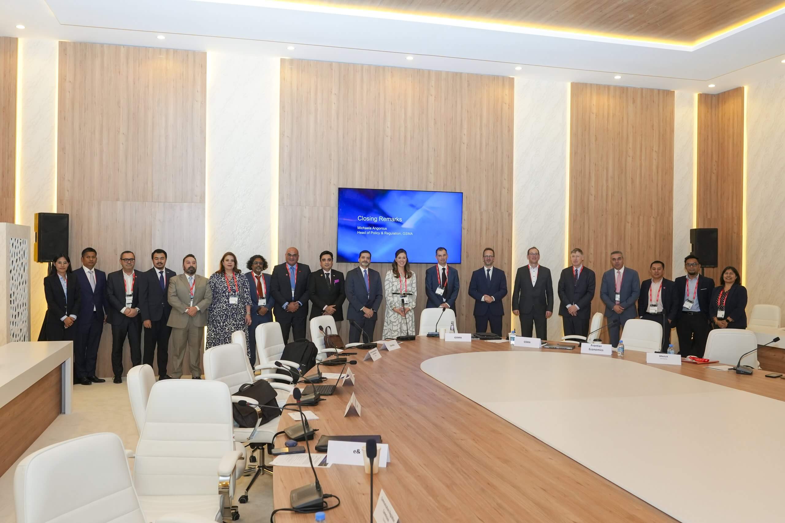 A group of 17 formally dressed people stand in a line against a modern wood-panelled conference room wall. In front of them is a curved conference table with white chairs and microphones. A large screen on the wall displays a blue welcome message.