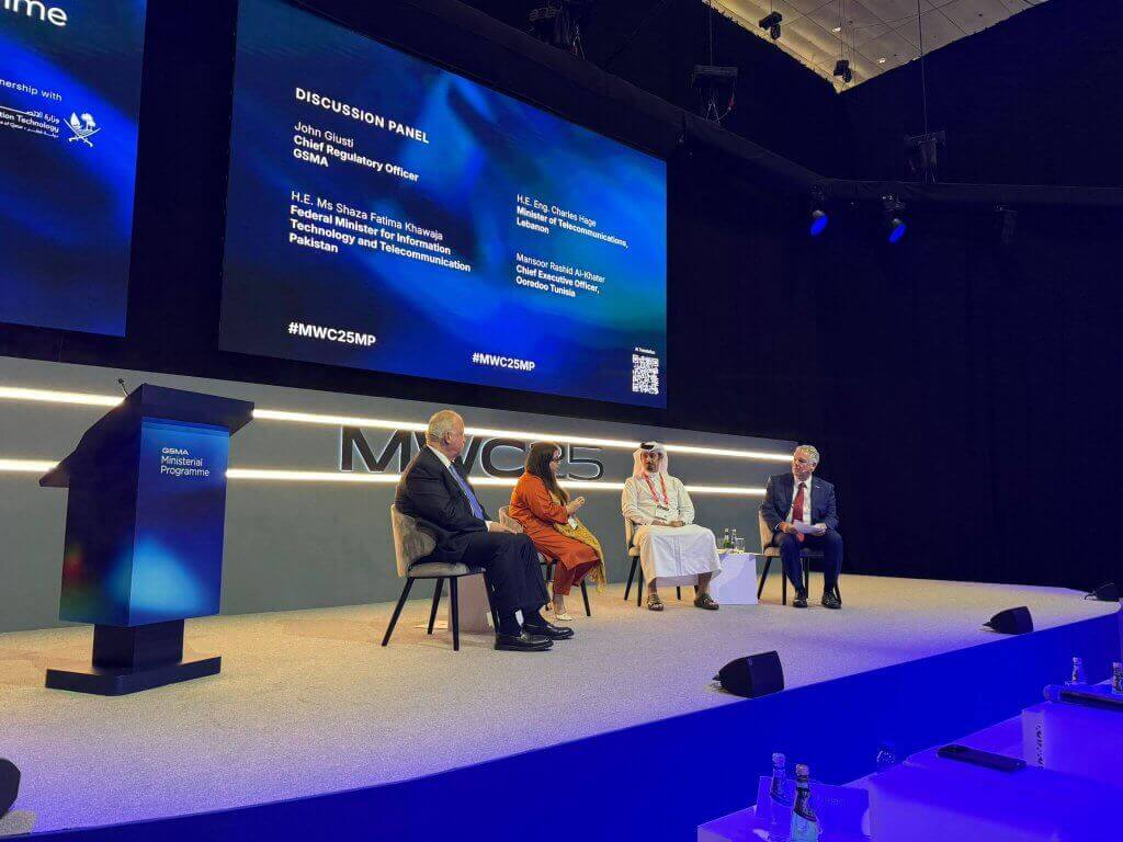 A panel of four people, three men and one woman, sit on stage in a modern conference room. Behind them, two large screens display panel details and names. The stage is well-lit, with "#MWC25MP" and "MWC25" visible on the displays and wall.