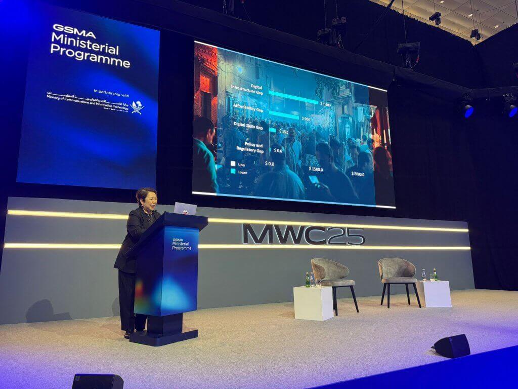 A speaker stands at a lectern labelled "GSMA Ministerial Programme" on a well-lit stage at MWC25. Behind them, a large screen displays event details and a digital crowd image. Two empty chairs and small tables with bottled water are on the stage.
