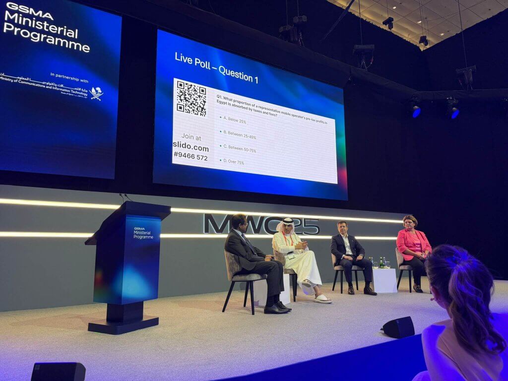 Four people sit onstage in front of a large screen displaying a live poll question at the GSMA Ministerial Programme Doha. The poll shows four answer options with bar graphs, set on a modern stage with blue lighting and a lectern to the left.