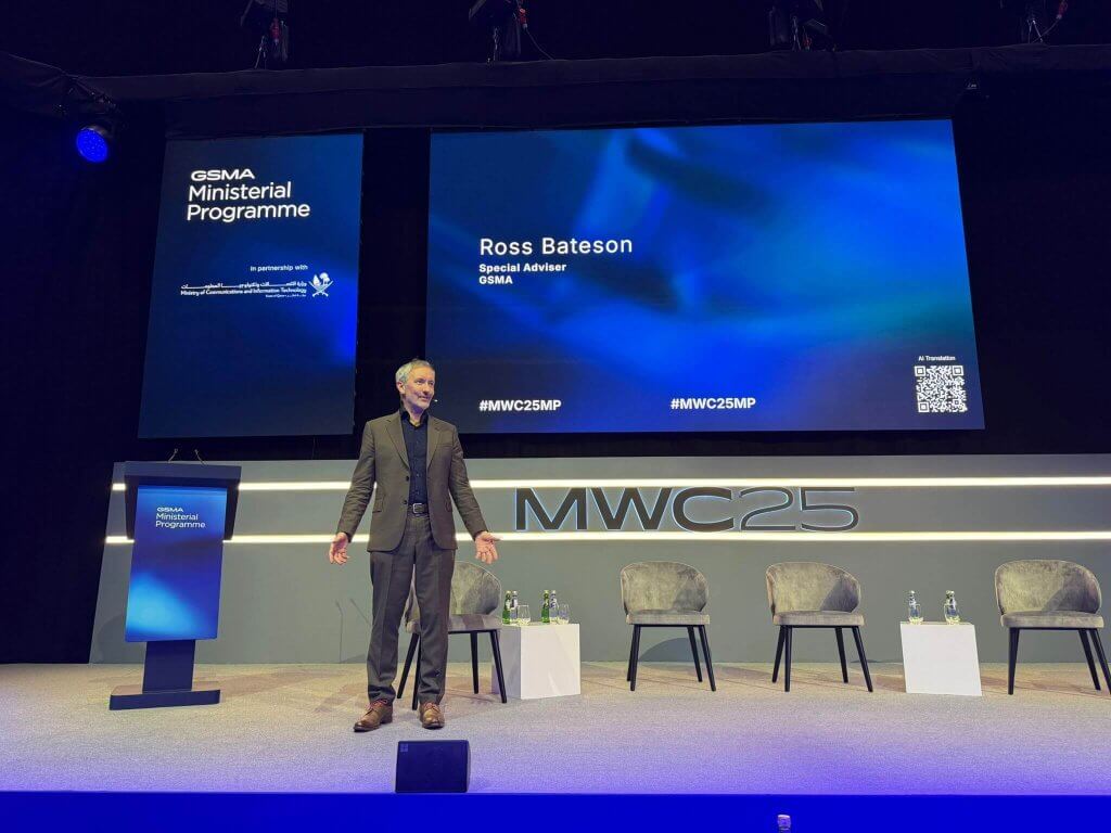 A man in a suit stands on stage smiling, with four empty chairs beside small tables holding water bottles. Behind him, a large screen displays “Ross Bateson, Special Adviser, GSMA” with hashtags #MWC25MP and a QR code. The stage backdrop is blue and reads “MWC25”.
