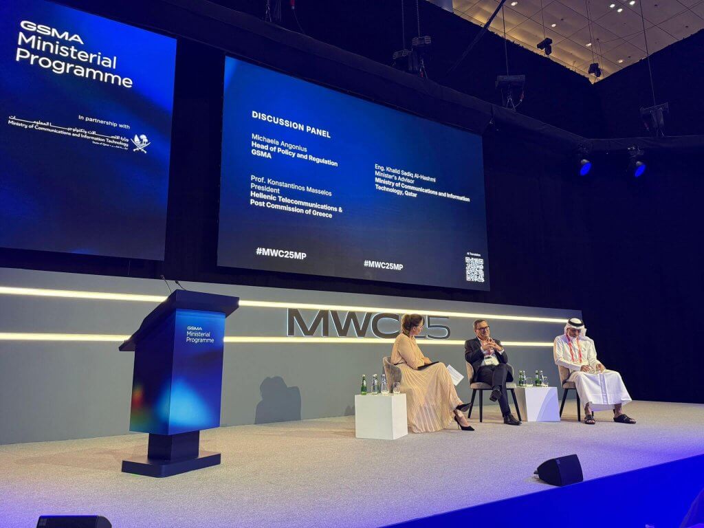 Three panellists sit on a stage with blue lighting at the Ministerial Programme Doha. Two men, one in a suit and one in traditional Gulf attire, and a woman in a green dress engage in discussion as large screens behind them display key details.