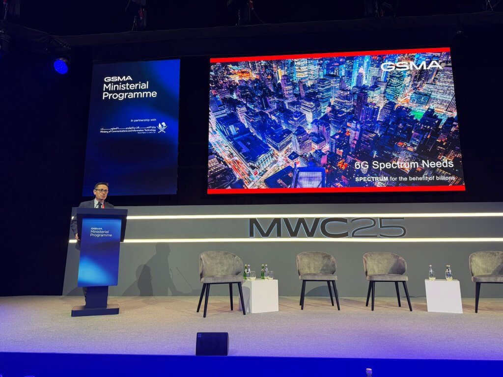 A man in a suit speaks at a podium onstage at the MWC25 event. Behind him, a screen shows a cityscape with the text “6G Spectrum Needs” and “GSMA.” Four empty chairs and water bottles are set up on stage. Blue and white lighting illuminate the scene.
