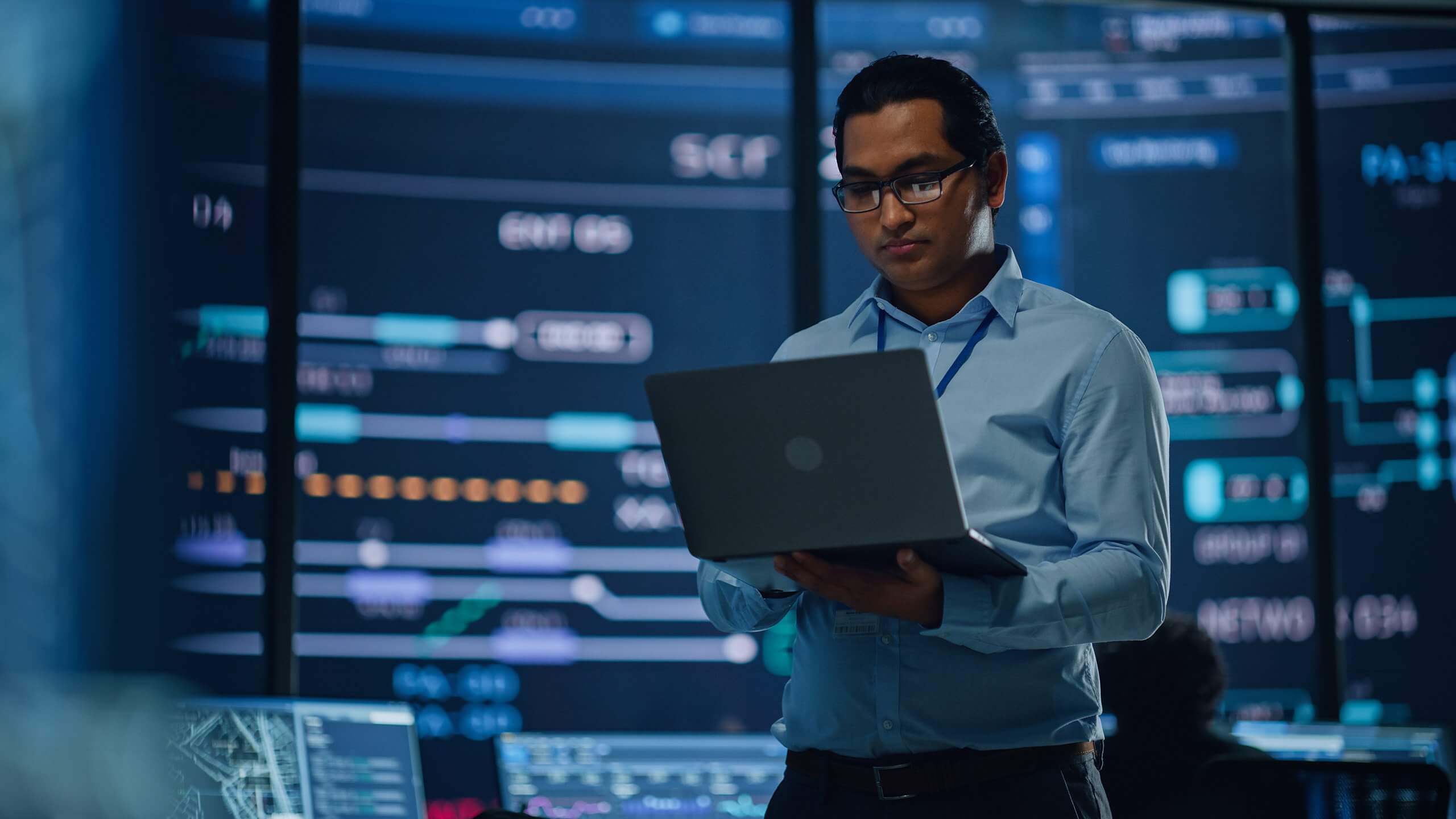 A man wearing glasses and a light blue shirt stands in a modern control room, holding a laptop. Multiple large digital screens display complex data, reflecting a high-tech, analytical environment focused on GSMA Public Policy initiatives.