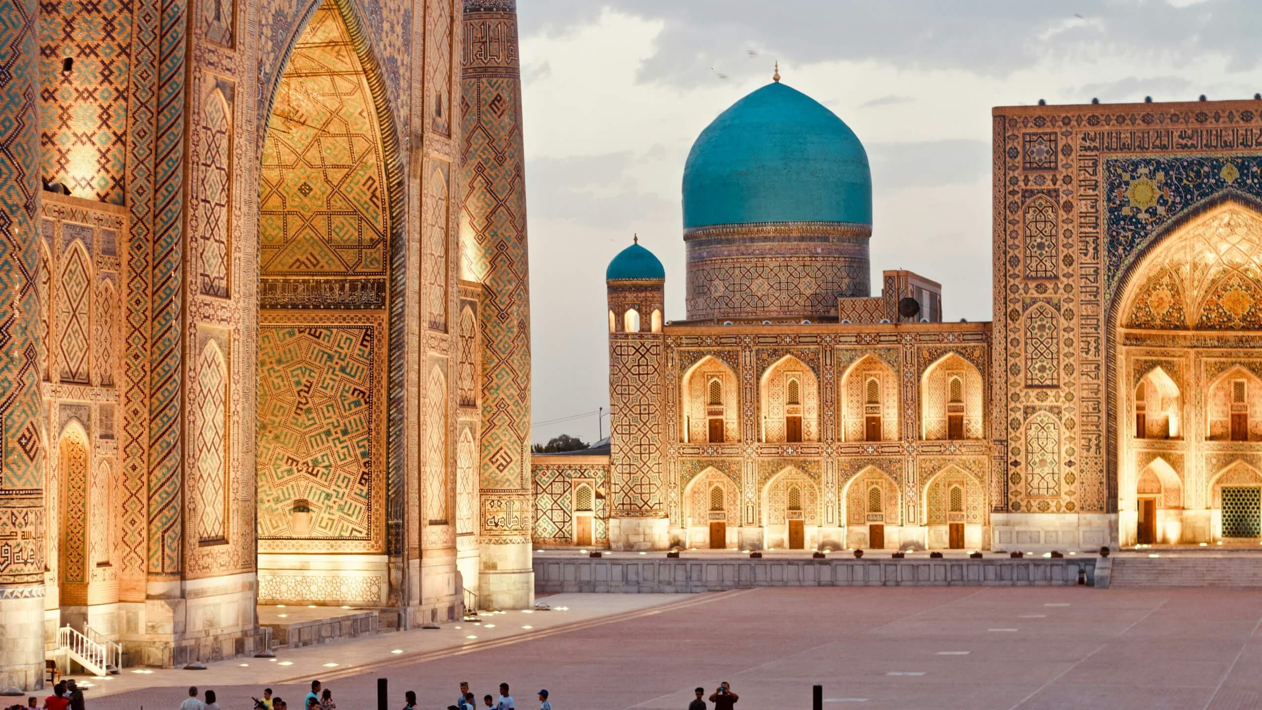 Historic square with ornate Islamic architecture; three large buildings with GSMA Public Policy-inspired intricate geometric tile patterns, two featuring huge arched entrances, and one topped with a turquoise dome. Evening light glows on the façades; small groups stroll in the open plaza.