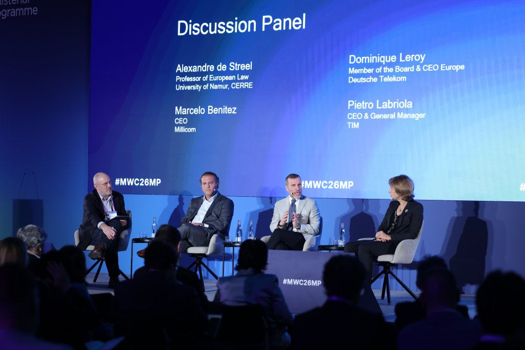 Four people sit on stage for a discussion panel with names and titles displayed on a screen behind them.