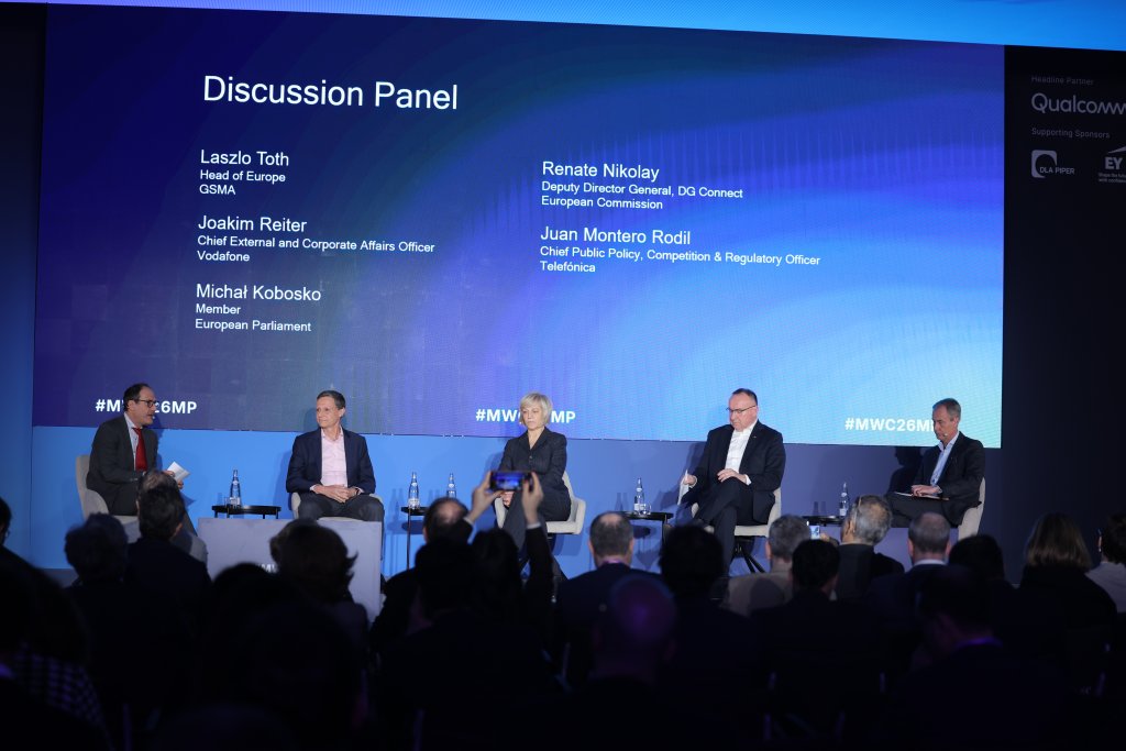 Four panelists and a moderator sit on stage discussing before a blue screen showing their names and titles.