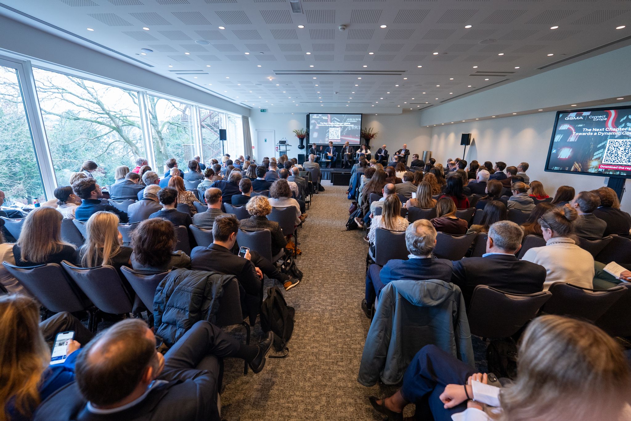 A large audience in a conference room watches a panel discussion at the front, with natural light pouring through big windows—an engaging scene typical of GSMA policy events.