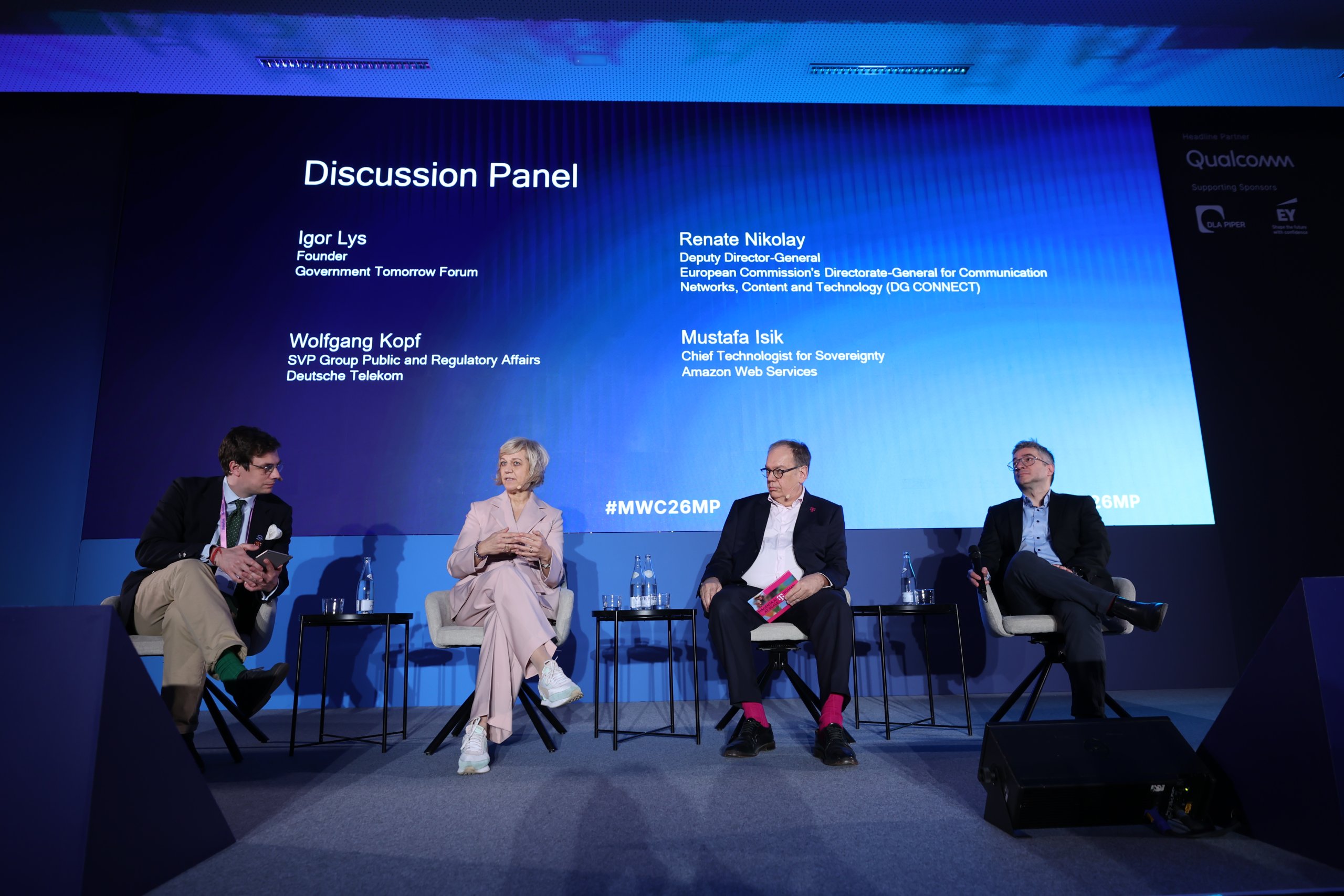 Four panelists sit on stage discussing, with their names and roles displayed on a blue screen behind them.