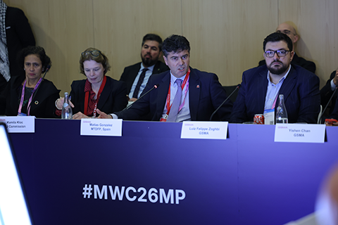 Four panelists sit at a conference table speaking at the #MWC26MP event, part of the GSMA policy events series, with nameplates in front of them.