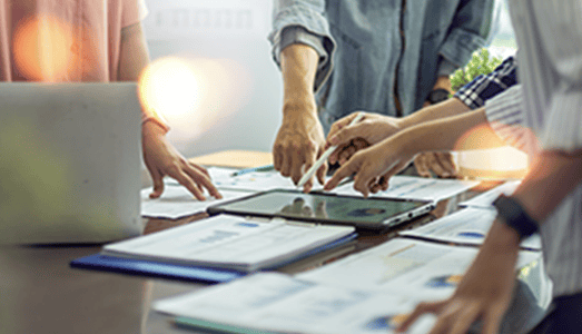 Four people pointing at papers and a tablet on a desk during a collaborative meeting or discussion.