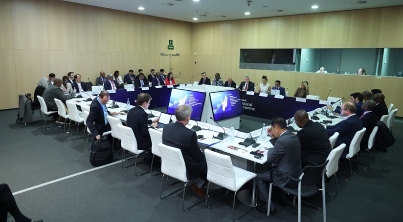 A group of people in business attire sit around a large conference table in a modern meeting room.