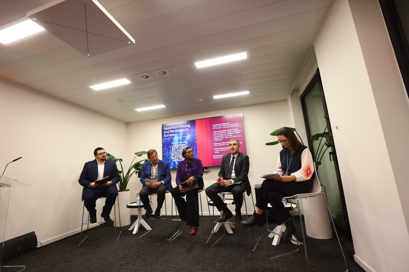 Five people sit on a panel in a modern office, discussing GSMA policy events in front of two digital display screens.