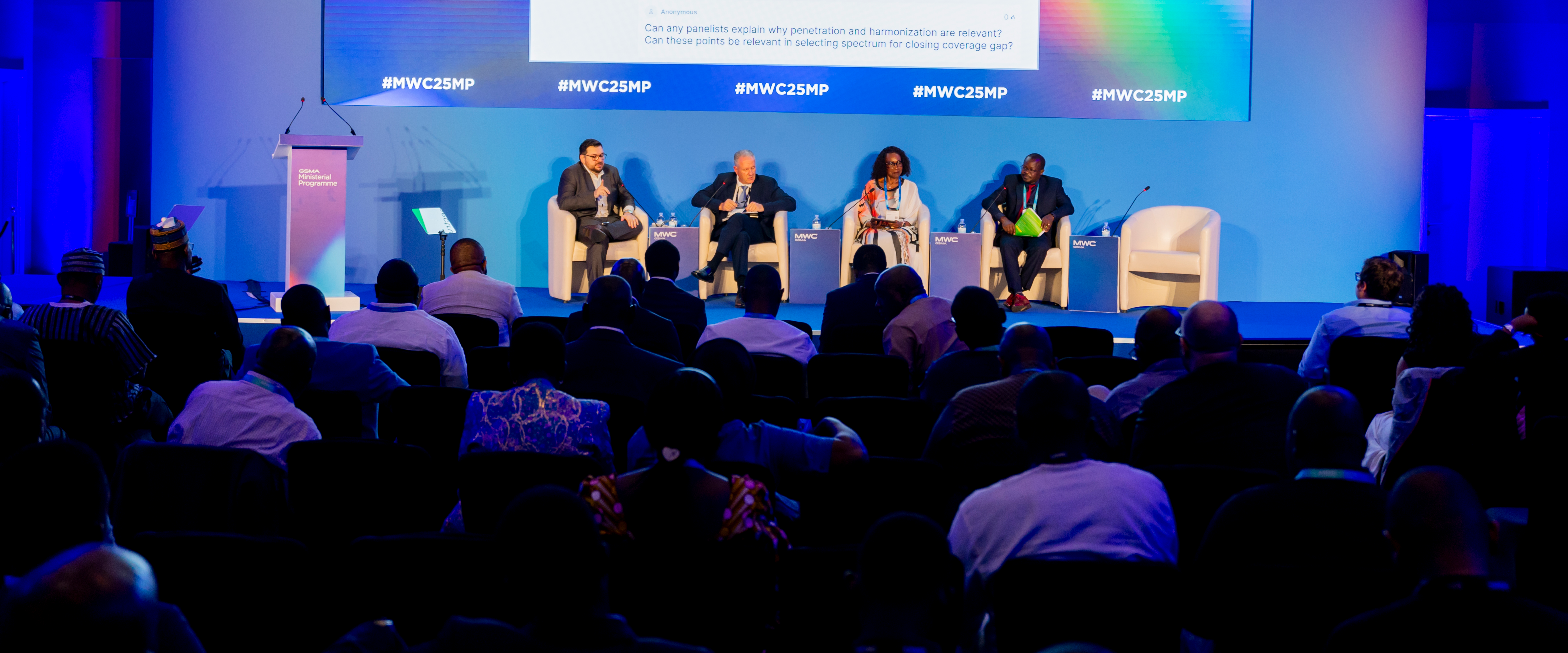Panelists sit on stage discussing at a GSMA policy event, with audience members watching in a blue-lit room.