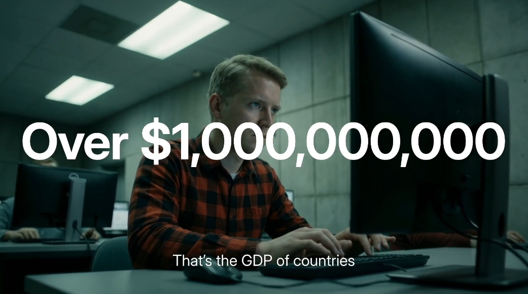 A man in a plaid shirt works at a computer. Text: "Over $1,000,000,000. That's the GDP of countries—and also the cost of scams each year.