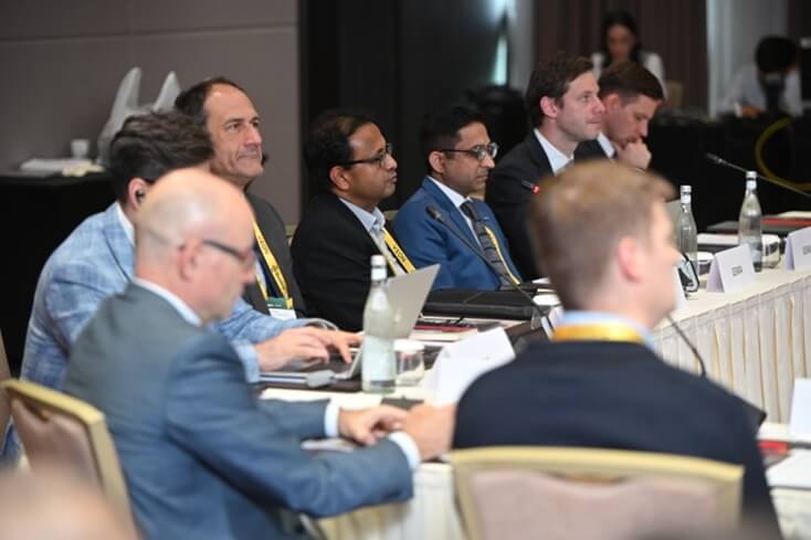 A group of people in business attire sit at a conference table, listening attentively during a meeting.