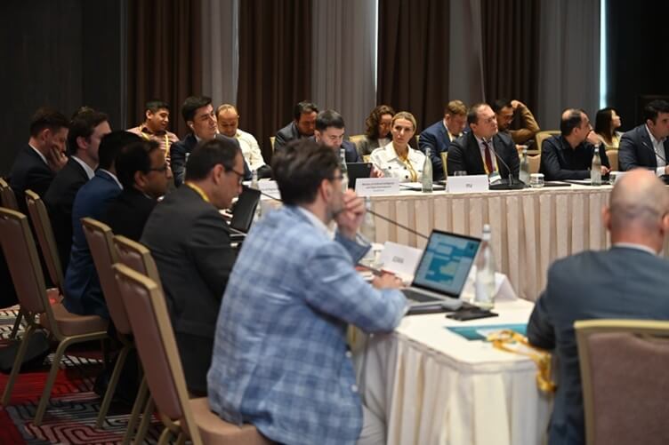 A group of people in business attire sit around a conference table with laptops and nameplates.