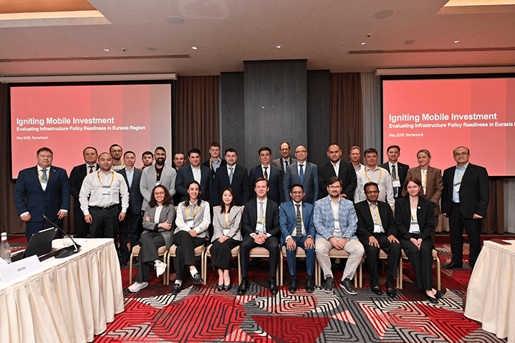 A group of people in business attire poses for a photo at a conference with presentation screens behind them.