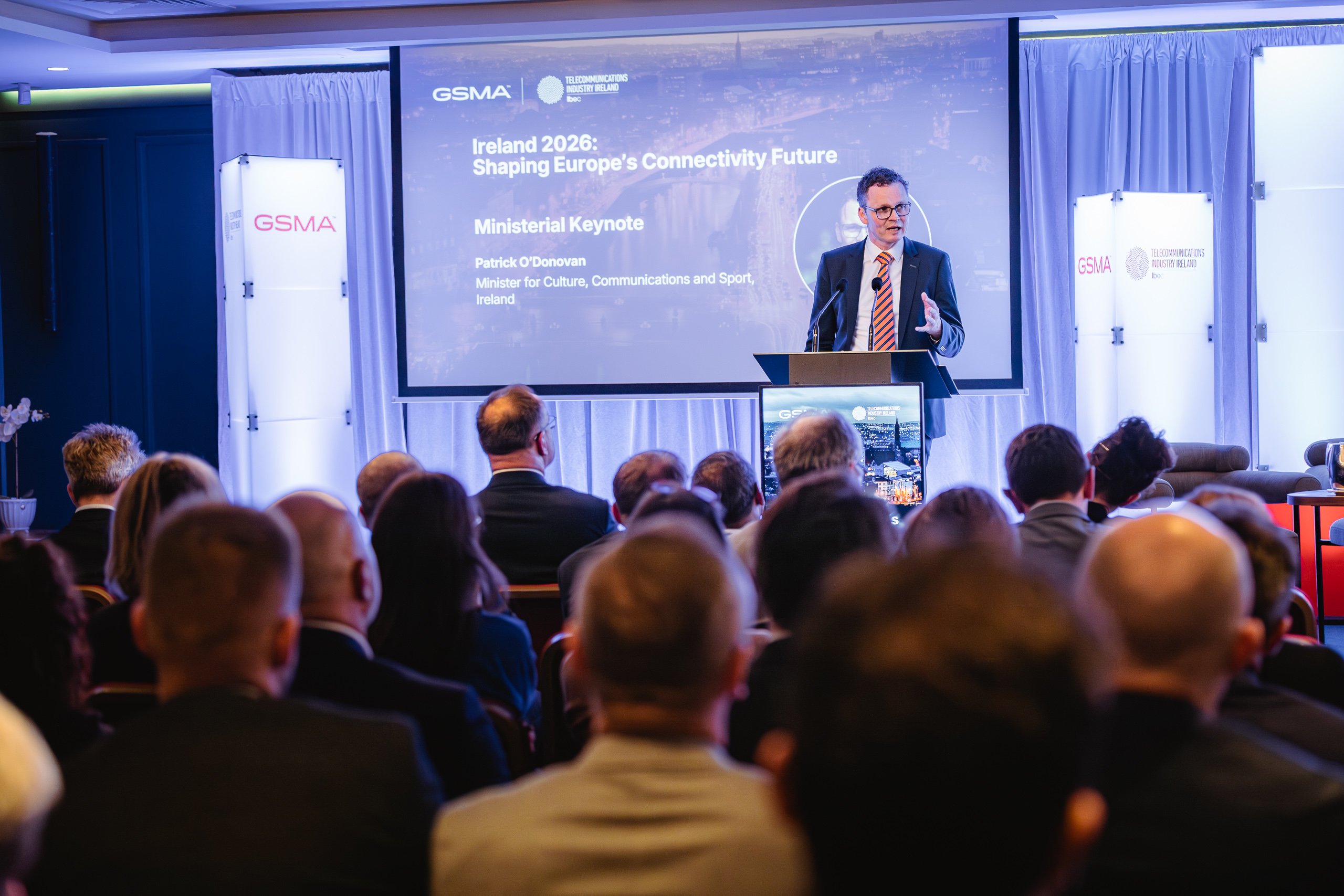 A man in a suit speaks at a podium to an audience during one of the prominent GSMA policy events. A screen behind him reads, "Ireland 2026: Shaping Europe’s Connectivity Future" with details about a ministerial keynote. The event appears formal, with seated attendees and professional lighting.