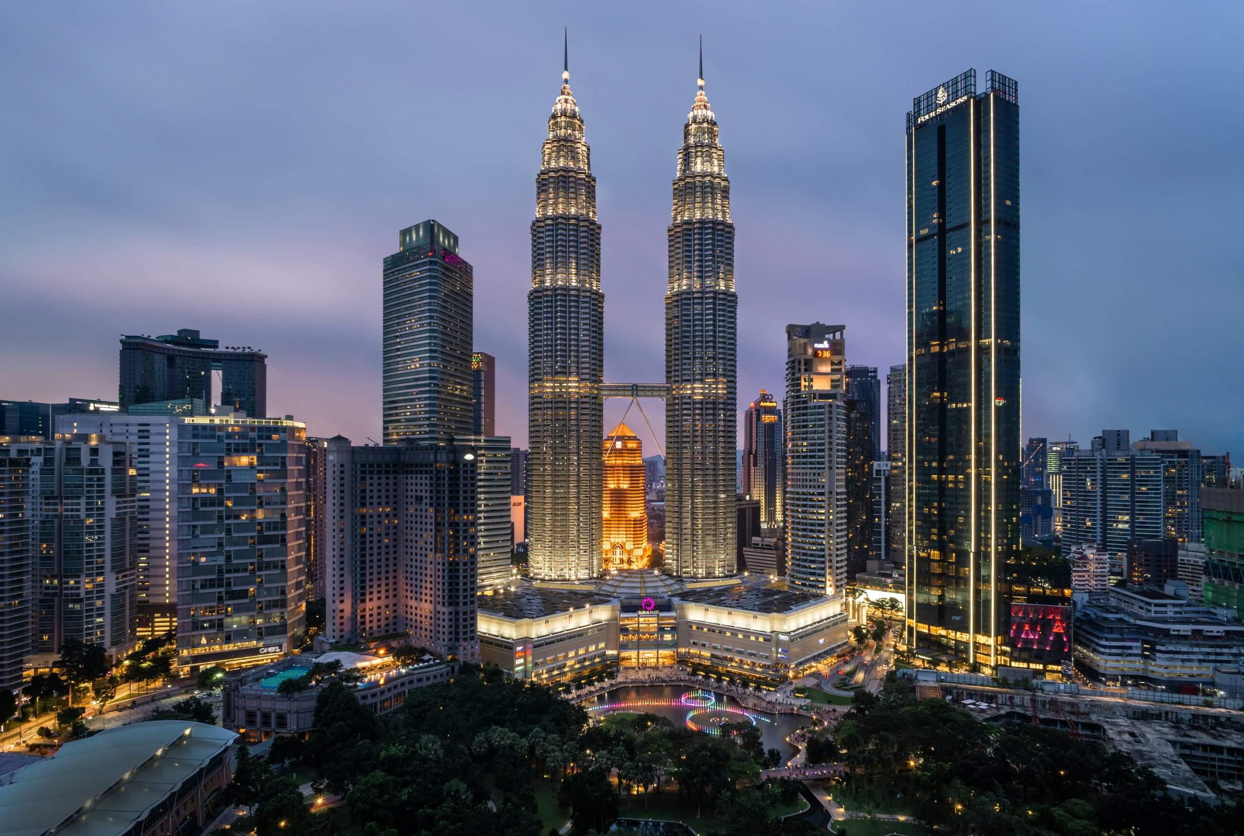 A cityscape at dusk shows the illuminated Petronas Twin Towers in Kuala Lumpur, a growing center for mobile industry public policy, surrounded by modern skyscrapers and busy streets framed by a park under a purple-blue sky.