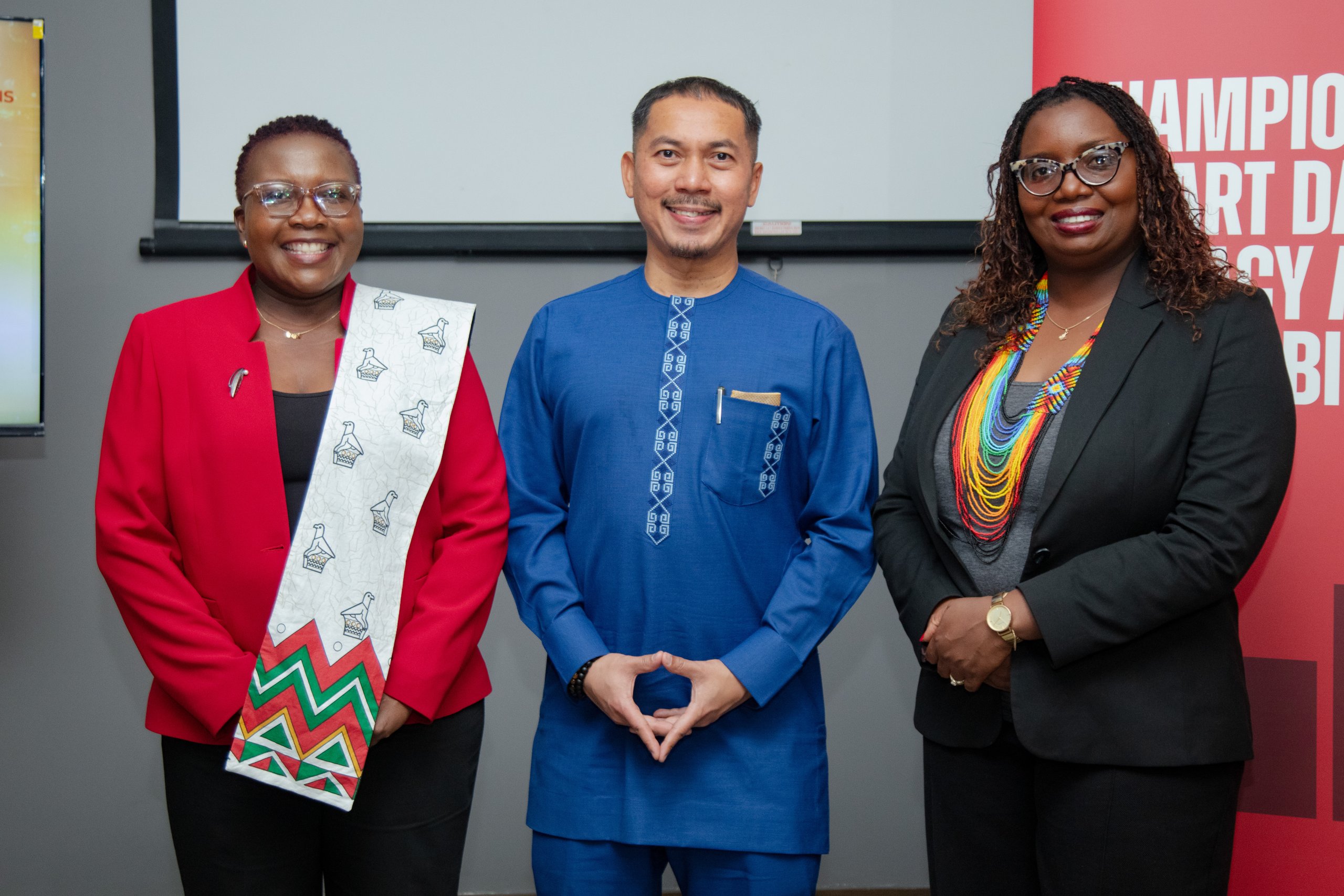 Three people stand side by side, smiling at the camera. The person on the left wears a red jacket and patterned sash, the center person wears a blue embroidered outfit, and the person on the right wears a black suit with a rainbow beaded necklace.